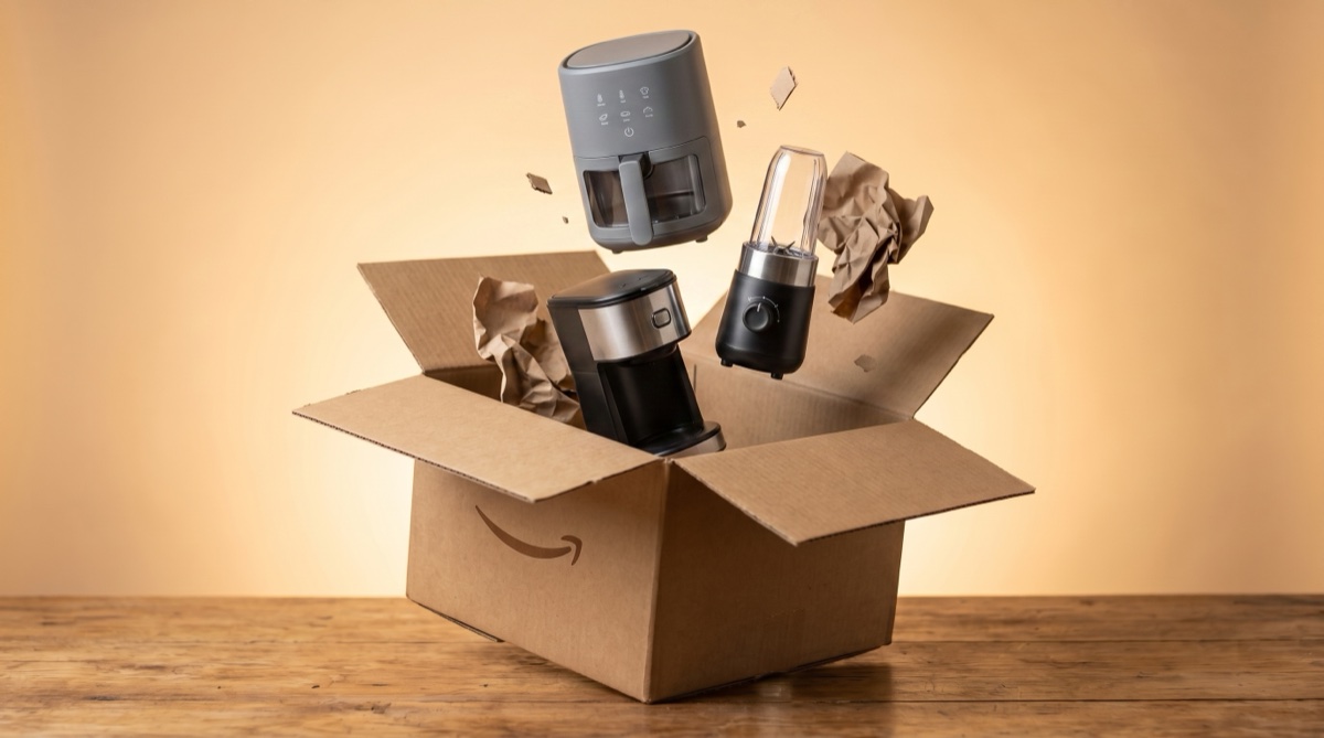 Uma caixa de entrega de papelão aberta com eletrodomésticos — air fryer, ventilador e micro-ondas — saindo dela e suspensos no ar, sobre fundo âmbar